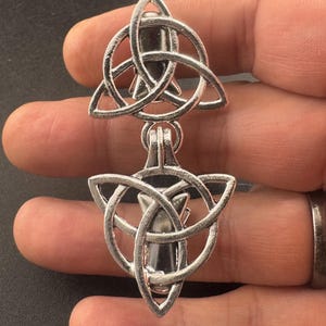 May include: A silver-toned metal brooch featuring two stacked triquetra symbols. Each triangular knot design is crafted from polished silver-coloured metal, with a dark, smooth, teardrop-shaped inlay centred within the interlocking loops of each knot.
