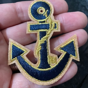 May include: An embroidered patch in the shape of a navy blue anchor with a gold rope wrapped around the centre. The anchor is outlined in gold thread and set against a light beige felt backing, creating a distinct nautical design element.