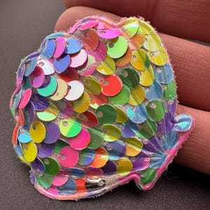 May include: A shell-shaped patch covered in iridescent, multi-coloured sequins in shades of pink, green, blue, yellow, and purple. The sequins are layered in a scale-like pattern, creating a reflective surface on a white fabric base.