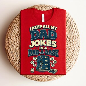 May include: A red t-shirt featuring the text I KEEP ALL MY DAD JOKES IN A DAD-A-BASE in blue and cream lettering. Below the text is a graphic of a stacked server tower with a smiling face, surrounded by speech bubbles containing the word HA.