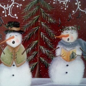 Puede incluir: Dos mu&ntilde;ecos de nieve con caras pintadas est&aacute;n cantando villancicos frente a un &aacute;rbol de hoja perenne nevado. El mu&ntilde;eco de nieve de la izquierda lleva un sombrero verde y un abrigo verde. El mu&ntilde;eco de nieve de la derecha lleva una bufanda azul y sostiene un libro.