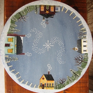 May include: A round wooden plate painted with a winter scene. The plate features four small houses with snow-covered roofs and chimneys. The houses are surrounded by trees and a swirling pattern of white dots that resemble falling snow. The plate is painted on a blue background with a snowflake in the center. The text "GENERAL STORE" is painted on the front of one of the houses.
