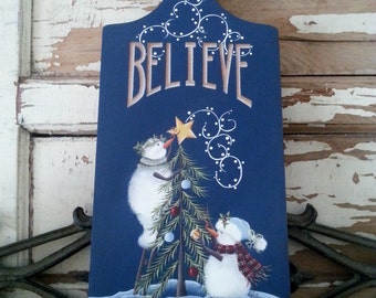 May include: A blue wooden cutting board with a white snowflake design at the top. The board has the word "BELIEVE" written in brown letters. Two snowmen are decorating a green Christmas tree with a yellow star on top. The snowmen are wearing red and white scarves.