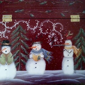 Op de afbeelding: Drie witte sneeuwpoppen met zwarte ogen en knopen, staand voor een rode achtergrond met witte sneeuwvlokken en krulletjes. De sneeuwpop links houdt een stok vast, de sneeuwpop in het midden houdt een boek vast en de sneeuwpop rechts speelt fluit.