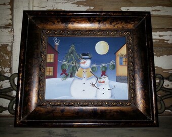 Puede incluir: Una pintura enmarcada de dos muñecos de nieve de pie frente a un paisaje nevado con casas y una luna llena. Los muñecos de nieve están decorados con adornos verdes y lazos rojos. La pintura tiene un marco marrón oscuro con detalles dorados.