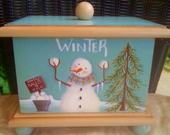 Puede incluir: Una caja de madera con una superficie pintada de azul claro y un diseño de muñeco de nieve. La caja tiene una tapa de madera con un pomo y las palabras "WINTER" pintadas en la parte delantera. El muñeco de nieve está sosteniendo bolas de nieve y tiene una bufanda y carámbanos.