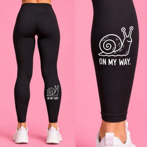 May include: Black leggings featuring a white line art graphic of a snail on the lower leg. Below the snail, the text reads ON MY WAY. The leggings have a high waistband and are shown from the back, paired with white athletic sneakers.