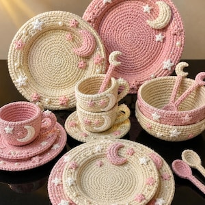 Op de afbeelding: Een set gehaakt servies bestaande uit borden, kopjes, schoteltjes en kommen in crème- en roze tinten. Elk item is versierd met kleine gehaakte sterretjes, maansikkels en witte kralen, voorzien van een gestructureerd, cirkelvormig steekpatroon.