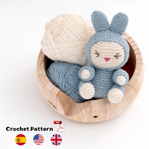 Puede incluir: Un pequeño muñeco de conejo tejido a ganchillo a mano con un traje azul con capucha, cara color crema y nariz rosa, sentado dentro de un cuenco de madera redondo junto a un ovillo de lana crema y otro azul. El texto Crochet Pattern es visible.