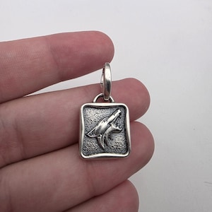 May include: A rectangular silver pendant featuring a raised relief of a howling wolf head on a textured background. The pendant has a smooth, rounded border and includes a silver jump ring at the top for attachment to a chain or cord.