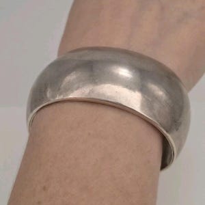 May include: A wide, polished silver-tone cuff bracelet with a smooth, rounded surface. The bracelet features a solid, continuous band design that wraps around the wrist, showcasing a reflective metallic finish and a simple, unadorned appearance.