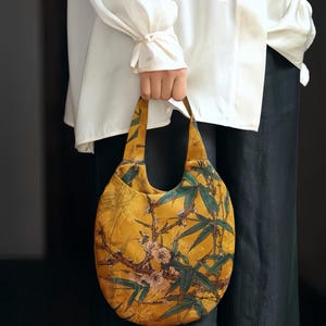May include: A round, mustard yellow handbag featuring a detailed print of green bamboo stalks and light pink cherry blossoms. The bag has a single integrated handle and a curved, circular silhouette, showcasing a nature-inspired botanical pattern.