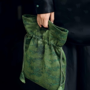 Può includere: Una borsa a sacchetto verde con coulisse caratterizzata da un motivo testurizzato a rami verde scuro su tutta la superficie. La borsa presenta un'apertura superiore arricciata con cordini verdi abbinati che pendono dai lati con linguette.