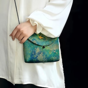 May include: A small crossbody bag featuring a mottled green and blue watercolor-style pattern with a yellow butterfly graphic on the front flap. The bag has a rounded flap closure secured by a single black bead and a thin black shoulder strap.