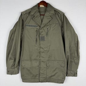 May include: An olive green field jacket featuring a collared neckline, long sleeves with buttoned cuffs, two vertical zippered chest pockets, and two large lower pockets with buttoned flaps. The jacket includes a rectangular hook-and-loop patch.