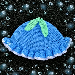 May include: A bright blue crocheted bucket hat featuring a ruffled white trim along the edge and two light green crocheted leaves attached to the top centre. The hat is constructed with a textured yarn stitch pattern throughout the main body.