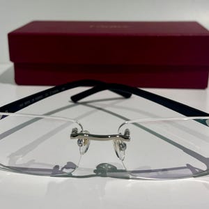 Puede incluir: Gafas sin montura con lentes rectangulares transparentes, un puente de metal plateado y patillas de color oscuro. Las gafas están colocadas frente a un estuche rectangular de color burdeos con un acabado liso y sólido.
