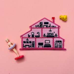 May include: Pink felt house patch with a black outline and interior details. The house has multiple floors and rooms with furniture and appliances. The patch is perfect for adding a touch of whimsy to clothing, bags, or other accessories.