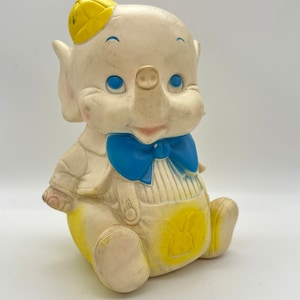 Peut inclure: Un éléphant en caoutchouc vintage en forme de jouet portant un chapeau jaune, un nœud papillon bleu et une salopette avec un motif de lapin jaune. Le jouet a un corps blanc et des yeux bleus.