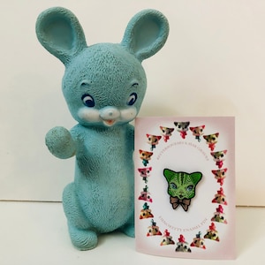 May include: A green and white enamel pin featuring a cat with a bow tie. The pin is surrounded by a circle of smaller cat illustrations. The text "BitterSqueaks & Mab Gravers" and "Dinkitty Enamel Pin" are printed on the card.
