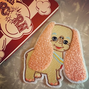 May include: A pink and beige felt applique of a dog with floppy ears. The dog has a blue and yellow striped collar and a smiling face.