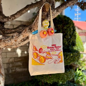Puede incluir: Un bolso de tela color crema con un estampado gráfico en rojo y amarillo de un cohete y un astronauta. El texto dice Rickey-Rocket PULL TOY FOR YOUNG ASTRONAUTS. Un pequeño juguete con ruedas naranjas está sujeto al asa superior del bolso.