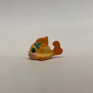 May include: A small yellow and orange plastic toy fish with a blue bow. The fish has a painted face with a blue eye and pink cheeks.