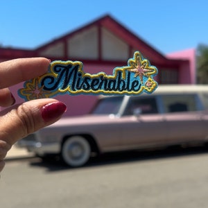 May include: A blue and yellow embroidered patch with the word "Miserable" in black lettering. The patch has two gold star accents.