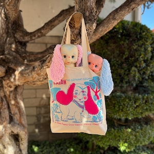 Könnte beinhalten: Eine beige Stofftasche mit einer Grafik eines Zeichentrick-Elefanten mit großen rosa Ohren. Der Text lautet RAJAH THE ELEPHANT ELEGANT und To Keep Teeny Ones Happy Happy Happy. Zwei Plüschelefanten mit rosa und blauen Ohren schauen oben heraus.