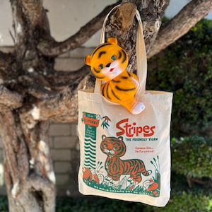 Puede incluir: Una bolsa de tela beige con un estampado gráfico de un tigre con el texto Stripes The Friendly Tiger, Squeeze him... He Purrrrrrs!, e It's an Arrow Toy. Un juguete de tigre de plástico moldeado se encuentra dentro de la bolsa.