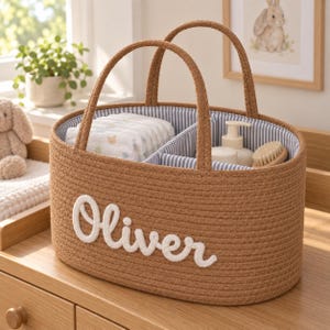 May include: An oval-shaped brown woven rope storage basket with two handles. The exterior features the name Oliver in white cursive lettering. The interior is lined with blue and white striped fabric and contains a divider holding nappies and bottles.