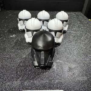 May include: A collection of six miniature helmets arranged in a triangular formation. Five helmets are white with black detailing, and one helmet in the front center is solid black. The helmets feature a smooth, molded design with distinct visor areas.