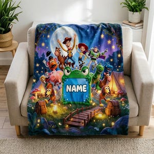 May include: A soft fleece blanket featuring a vibrant nighttime camping scene with Toy Story characters including Woody, Buzz Lightyear, Jessie, and Rex. A central green frog-shaped frame displays the text NAME above a wooden bridge.