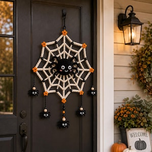 May include: A crocheted spider web wall hanging featuring a large central black spider with white eyes and a smiling mouth. The web is off-white with orange flower accents, black beads, and four smaller dangling black spiders at the bottom.