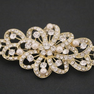 Puede incluir: Una pinza para el pelo dorada con un diseño floral. La pinza está adornada con diamantes de imitación transparentes y perlas falsas.