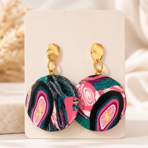 May include: A pair of dangle earrings featuring circular pendants with a marbled pattern in shades of black, hot pink, teal, and white. The pendants are attached to gold-toned metal studs with a hammered texture, connected by small gold-toned jump rings.