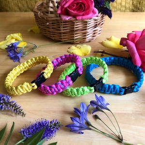 May include: Four paracord bracelets featuring a woven pattern and black plastic side-release buckles. The bracelets are coloured yellow, pink and purple, lime green, and bright blue, arranged in a row on a light wooden surface.