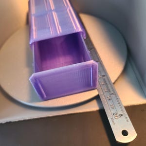 May include: A purple 3D-printed rectangular storage box with an open drawer, positioned next to a metal ruler showing a length of approximately 20 cm. The object features a layered texture and a hollow interior compartment for storage.