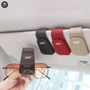 Könnte beinhalten: Vier Sonnenbrillenhalter für die Sonnenblende im Auto aus lederartigem Material in Schwarz, Rot, Beige und Dunkelbraun. Jeder Halter verfügt über einen Magnetverschluss und das weiße STI-Logo, das auf der Vorderseite aufgedruckt ist.