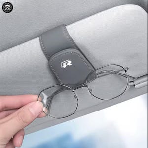 Könnte beinhalten: Ein grauer Brillenhalter aus Leder für die Sonnenblende im Auto mit einem weißen R-Logo. Der Halter verfügt über einen Magnetverschluss und ist so konzipiert, dass er an der Sonnenblende befestigt wird, hier mit einer sechseckigen Brille.