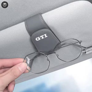 May include: A grey leather car visor sunglasses holder featuring the white text GTI printed on the front. The clip is attached to a light grey car sun visor and holds a pair of thin-framed metal eyeglasses securely in place with a magnetic closure.