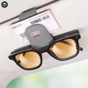 Op de afbeelding: Een grijze zonneklepclip met het Audi-logo in wit en rood, die een zonnebril met zwart montuur en amberkleurige glazen vasthoudt. Een witte rechthoekige kaart met een streepjescode en tekst zit achter de clip op de zonneklep geklemd.