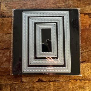 May include: A set of four nested silver metal rectangular cutting dies with a stitched border pattern. The dies are arranged concentrically on a black backing sheet and enclosed within a clear plastic sleeve for storage and protection.