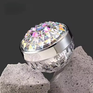 May include: A round, clear acrylic container with a faceted, diamond-like texture on the base and a lid featuring a cluster of iridescent, multi-colored, pyramid-shaped crystals that reflect light in shades of pink, blue, and yellow.