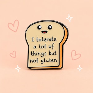 May include: A cream-coloured enamel pin shaped like a slice of bread with a black outline. The bread has a smiling face with two black eyes and a small mouth. The text reads: I tolerate a lot of things but not gluten. The side is orange.