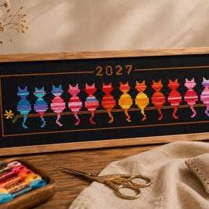 Pode incluir: Um bordado em ponto cruz retangular numa moldura de madeira clara com doze gatos coloridos sentados numa linha horizontal sobre um fundo preto, com o ano 2027 bordado a laranja no centro superior e pequenas flores amarelas nas extremidades.