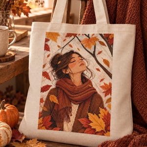 Puede incluir: Un bolso de tela beige con una ilustración impresa de una persona con el pelo recogido en un moño, vistiendo una bufanda y un suéter marrón grueso, rodeada de hojas de otoño que caen en tonos naranja, rojo y amarillo sobre un fondo blanco.