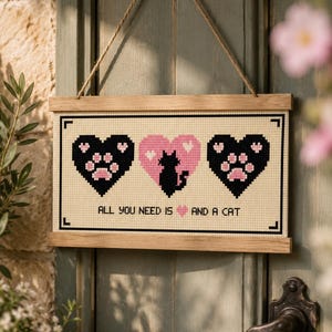 Puede incluir: Un tapiz rectangular de punto de cruz con dos corazones negros con huellas rosas y un corazón rosa central con la silueta de un gato negro. El texto dice ALL YOU NEED IS [corazón rosa] AND A CAT. Está enmarcado con listones de madera clara y colgado con cuerda.