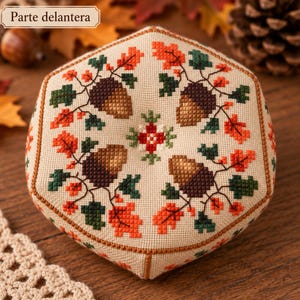 Könnte beinhalten: Ein sechseckiges Nadelkissen mit Kreuzstichmuster auf beigem Stoff. Das Design zeigt vier braune Eicheln, umgeben von orangefarbenen und dunkelgrünen Eichenblättern, mit einem kleinen rot-grünen Blumenmotiv in der Mitte.