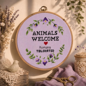 Puede incluir: Un bastidor de bordado circular de madera con tela color lavanda y un diseño de punto de cruz. El texto dice ANIMALS WELCOME y HUMANA TOLERATED en negro, separados por un corazón rojo, rodeados por una corona de hojas verdes y flores moradas.